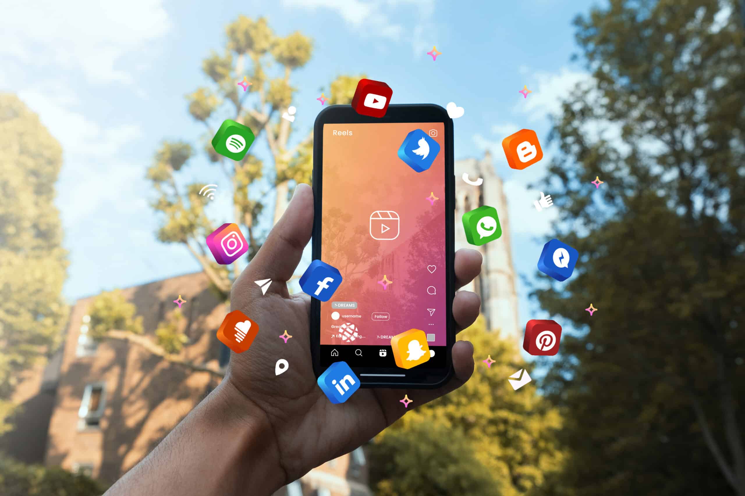 Image d'une main qui tient un smartphone affichant une interface de réseaux sociaux, avec des icônes de diverses plateformes flottant autour, sur un fond extérieur ensoleillé.